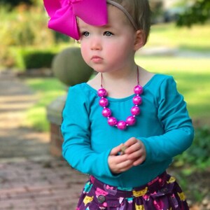 May include: A young girl wearing a turquoise long-sleeved shirt, a purple skirt with a cat print, a pink beaded necklace, and a large pink bow headband.