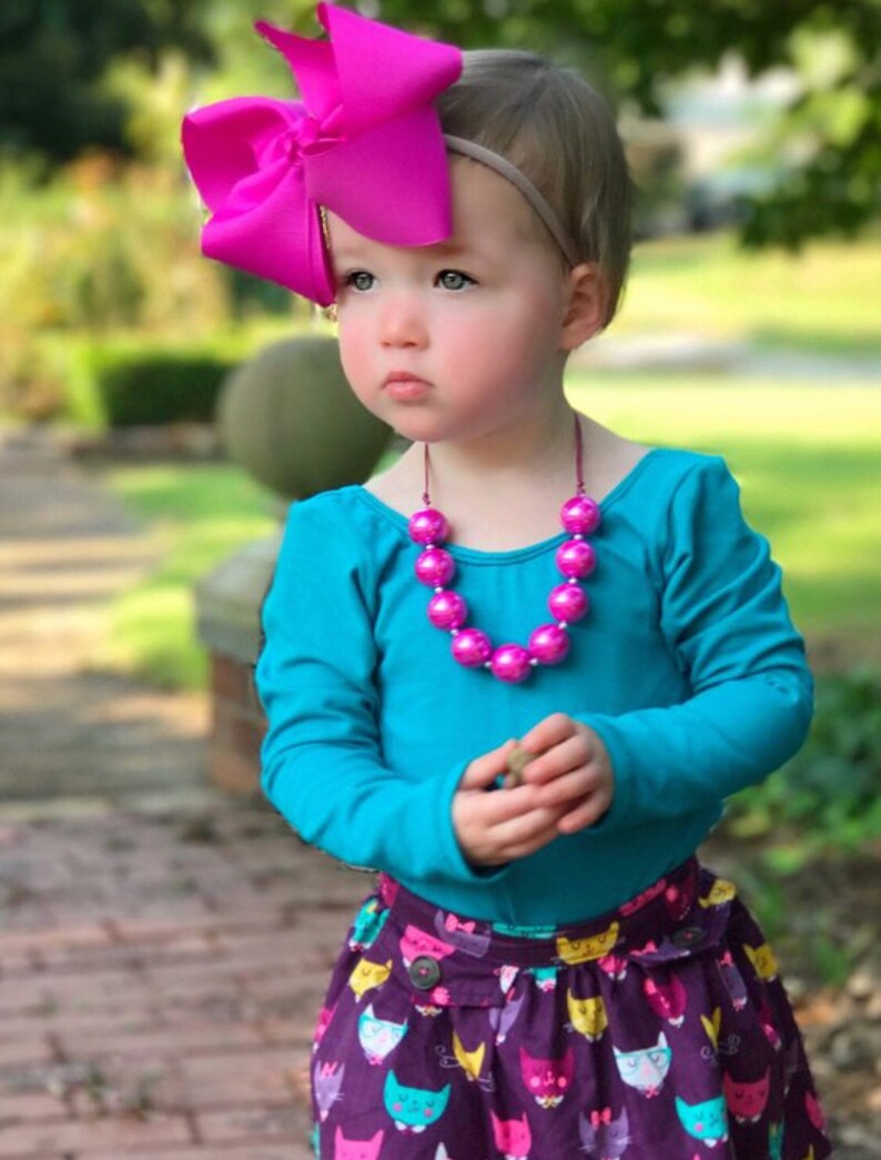 May include: A young girl wearing a turquoise long-sleeved shirt, a purple skirt with a cat print, a pink beaded necklace, and a large pink bow headband.