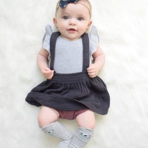 May include: A baby wearing a gray sleeveless bodysuit with black suspenders, a dark gray skirt, and gray knee-high socks with a white eye design. The baby is wearing a black and white polka dot bow headband.