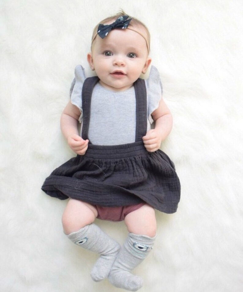 May include: A baby wearing a gray sleeveless bodysuit with black suspenders, a dark gray skirt, and gray knee-high socks with a white eye design. The baby is wearing a black and white polka dot bow headband.