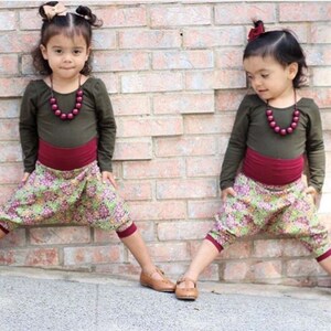 May include: Two young girls wearing matching outfits. They are both wearing a long-sleeved olive green shirt with a maroon waistband and floral print pants. They are both wearing a maroon beaded necklace and have their hair pulled back with a bow.