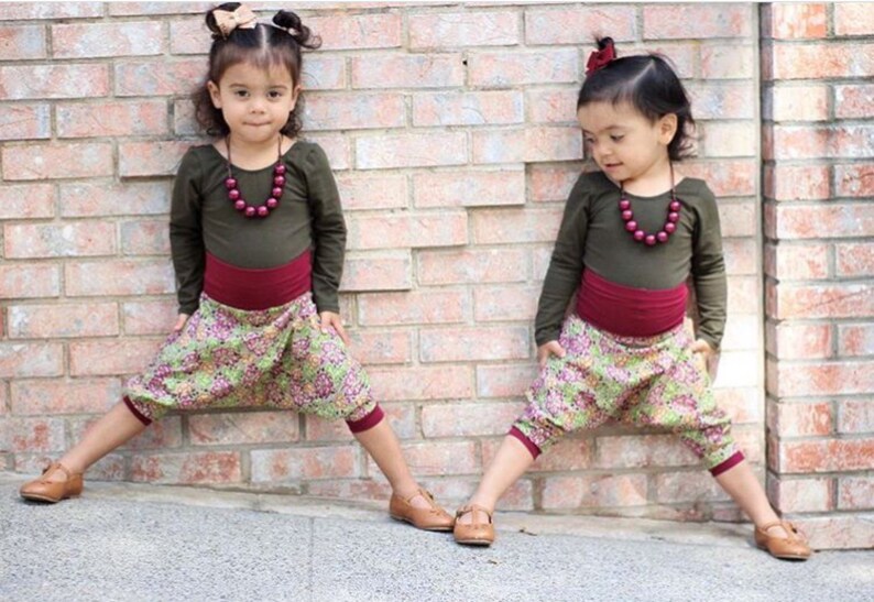 May include: Two young girls wearing matching outfits. They are both wearing a long-sleeved olive green shirt with a maroon waistband and floral print pants. They are both wearing a maroon beaded necklace and have their hair pulled back with a bow.