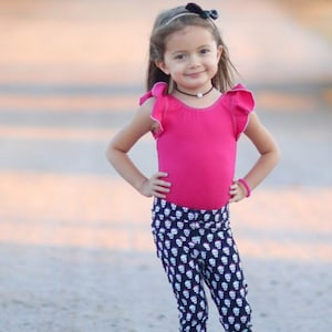 May include: A young girl wearing a pink leotard with ruffled sleeves and black leggings with a white skull pattern. She is wearing a black headband with a bow and a pearl necklace.