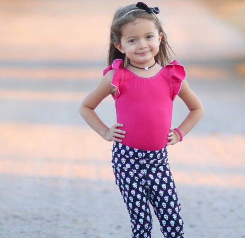 May include: A young girl wearing a pink leotard with ruffled sleeves and black leggings with a white skull pattern. She is wearing a black headband with a bow and a pearl necklace.