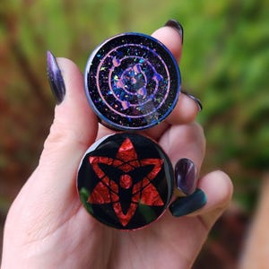 May include: Two round resin earrings with black backgrounds and colorful designs. The top earring has a purple and pink swirling design with a black outline. The bottom earring has a red star with a black outline.