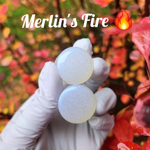 Puede incluir: Dos expansores de oreja blancos iridiscentes con un acabado brillante. Los expansores se sostienen en una mano enguantada. El texto "Merlin's Fire" es visible en la imagen.
