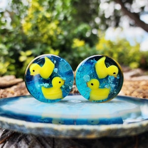 Könnte beinhalten: Zwei runde, transparente Glas-Ohrstecker mit gelben Enten auf blauem Hintergrund. Der Hintergrund ist verschwommen, grün und blau.