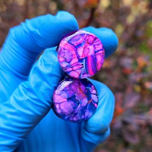 May include: Two round, purple and pink plugs with a marbled design. The plugs are held by a person wearing blue gloves. The background is blurred, with hints of green and brown.