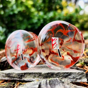 May include: Pair of clear glass ear gauges with red and white swirl patterns. The gauges feature a small silver and red knife design. The gauges are 1 3/8 inches (45 mm) in diameter.