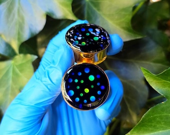 May include: Two gold-coloured ear gauges with a black face, patterned with blue, green, and yellow dots. The gauges are held against a backdrop of green ivy leaves.