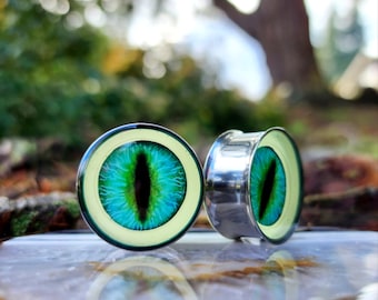 May include: A pair of green eye-shaped ear gauges with a silver metal rim. The gauges are on a light brown surface.