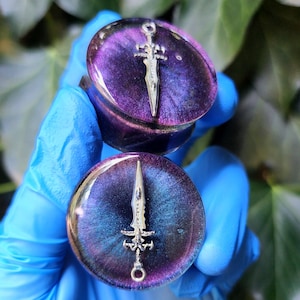 May include: Two purple and blue ear gauges with a silver sword design. The gauges have a glossy finish and a swirling color pattern. The sword is detailed with a hilt and blade. The background is blurred with green foliage.