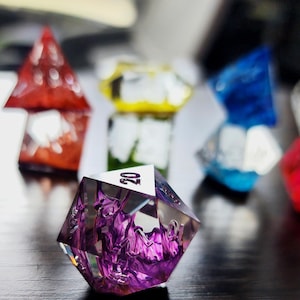 May include: A set of colorful, clear, resin dice with a purple 20-sided die in the foreground. The dice are translucent and have a swirling, marbled effect.