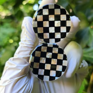 May include: Two round, black and white checkered earrings. The earrings have a glossy finish and are held up against a blurred green background. The checkered pattern is made up of alternating black and white squares.