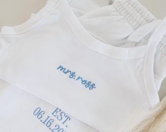 May include: White pajama set with blue embroidered text. The top says "mrs. ross" and the bottom says "EST. 06.16.2026". The set includes a tank top and shorts with a drawstring waist. A sprig of dried ornamental grass is in the foreground.