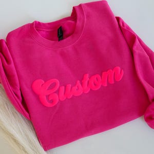 May include: A bright pink sweatshirt with the word "Custom" in a matching, raised font. The long sleeves are gathered at the wrists. The sweatshirt is made of a soft material and is suitable for casual wear.