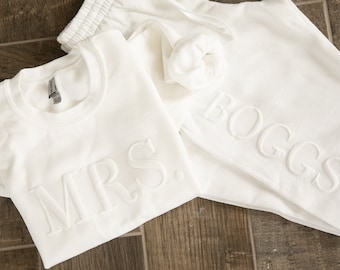 May include: White sweatshirt with the word "MRS." embroidered on the front and white sweatpants with the word "BOGGS" embroidered on the leg.
