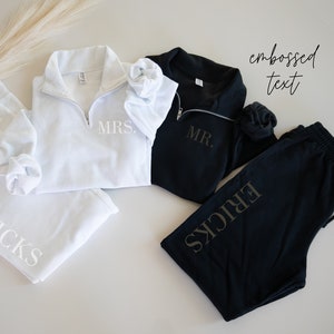 May include: White and black matching loungewear sets. The white set includes a zip-up sweatshirt with "MRS." and sweatpants with "ERICKS" printed on them. The black set includes a zip-up sweatshirt with "MR." and sweatpants with "ERICKS".