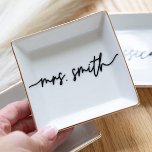 May include: White square ceramic trinket dish with gold trim, featuring the script text "mrs. smith" in black. The dish is held in a hand, with another similar dish visible in the background. The dish is likely used for jewelry or small items.