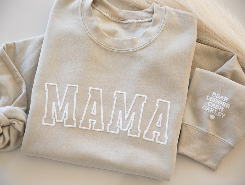 May include: A light beige sweatshirt with the word "MAMA" embroidered in white on the front. The sleeve has the names "BEAR, LEDGER, CASH, OAKLEY" embroidered in white.