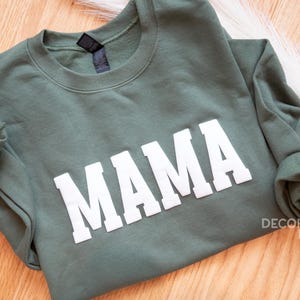 Puede incluir: Una sudadera verde con la palabra "MAMA" impresa en blanco en la parte delantera.