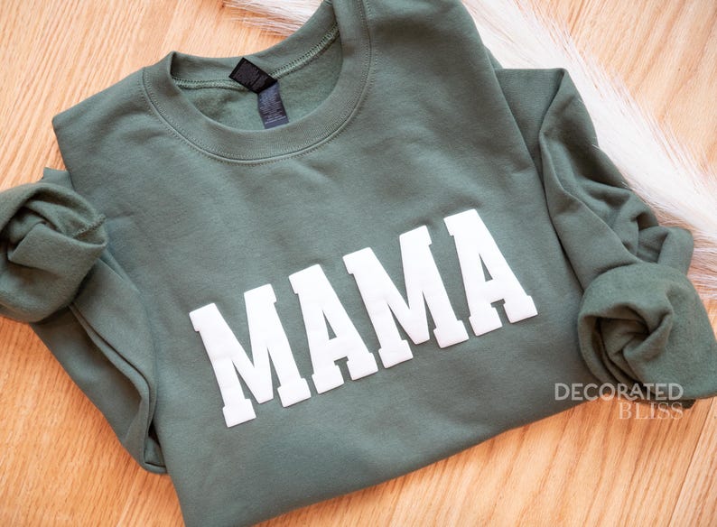 Puede incluir: Una sudadera verde con la palabra "MAMA" impresa en blanco en la parte delantera.