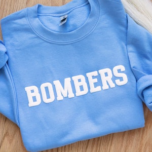 Puede incluir: Una sudadera azul claro con la palabra "BOMBERS" impresa en blanco en la parte delantera.