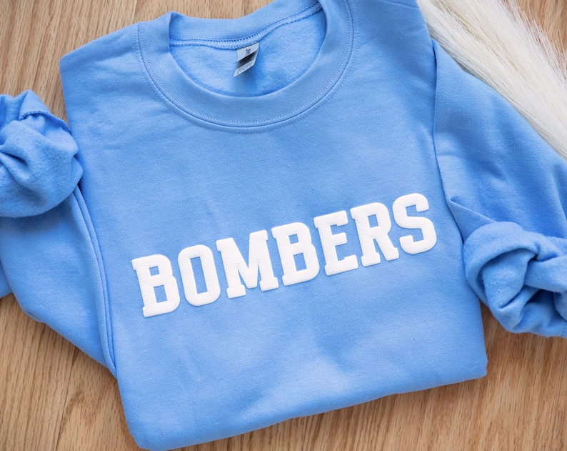 Puede incluir: Una sudadera azul claro con la palabra "BOMBERS" impresa en blanco en la parte delantera.