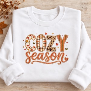 May include: A white long-sleeve sweatshirt featuring the text "COZY season" in a multi-patterned design. The letters in "COZY" display floral, plaid, leopard, and striped prints, while "season" is written in a solid orange cursive font.