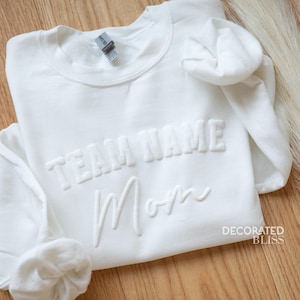 May include: White sweatshirt with the text "TEAM NAME Mom" in raised lettering. The sweatshirt is folded and the brand name "DECORATED BLISS" is visible in the bottom right corner.