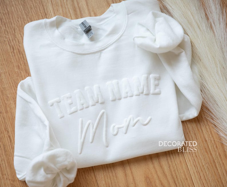 Puede incluir: Sudadera blanca con el texto "TEAM NAME Mom" en letras en relieve. La sudadera est&aacute; doblada y el nombre de la marca "DECORATED BLISS" es visible en la esquina inferior derecha.