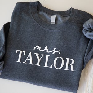 K&ouml;nnte beinhalten: Ein dunkelgraues Sweatshirt mit dem wei&szlig;en Schriftzug "mrs. TAYLOR". Das Sweatshirt ist gefaltet, ein &Auml;rmel ist sichtbar. Der Stoff wirkt weich und bequem, geeignet f&uuml;r Freizeitkleidung.