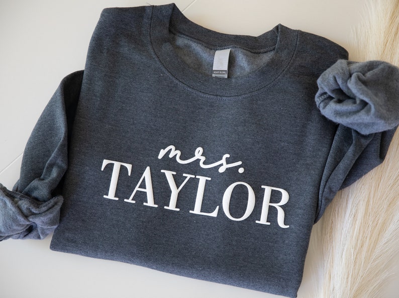 K&ouml;nnte beinhalten: Ein dunkelgraues Sweatshirt mit dem wei&szlig;en Schriftzug "mrs. TAYLOR". Das Sweatshirt ist gefaltet, ein &Auml;rmel ist sichtbar. Der Stoff wirkt weich und bequem, geeignet f&uuml;r Freizeitkleidung.