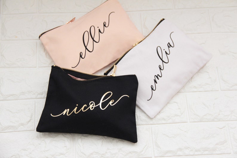 Puede incluir: Tres bolsas de maquillaje personalizadas con cierres de cremallera. Las bolsas son de color rosa, blanco y negro con letras doradas. Los nombres "ellie", "emelia" y "nicole" est&aacute;n escritos en cursiva en las bolsas.