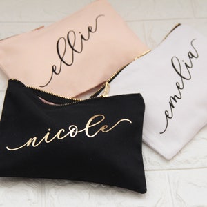 Puede incluir: Tres bolsas de maquillaje personalizadas con diferentes nombres. Las bolsas son negras, blancas y rosas. Los nombres est&aacute;n escritos en dorado y negro.