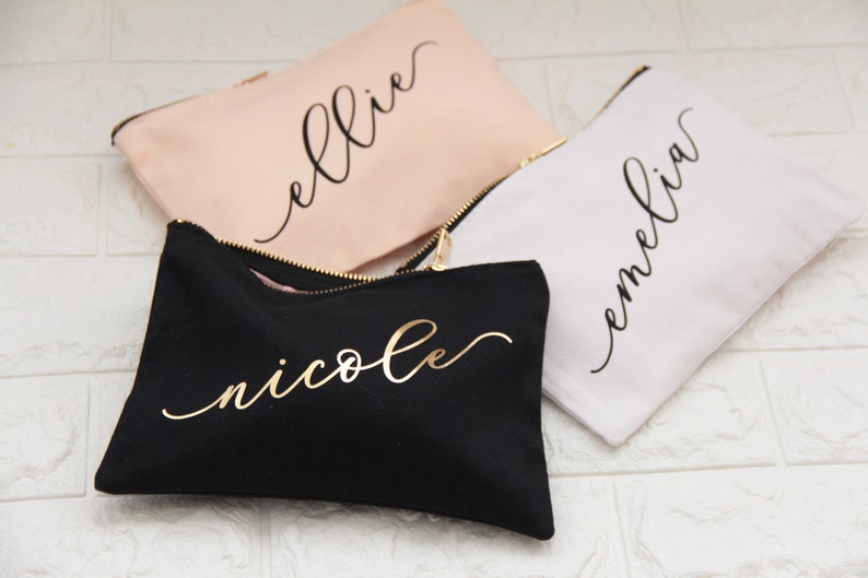 Puede incluir: Tres bolsas de maquillaje personalizadas con diferentes nombres. Las bolsas son negras, blancas y rosas. Los nombres est&aacute;n escritos en dorado y negro.
