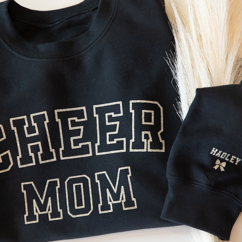 May include: Black sweatshirt with the words "CHEER MOM" in gold glitter lettering. The sweatshirt has a black sleeve with the name "HADLEY" and a small bow in gold glitter.