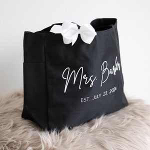 May include: Black canvas tote bag with white text that reads "Mrs.  Barber EST. JULY 23, 2024". A white bow is tied to the handle of the bag.