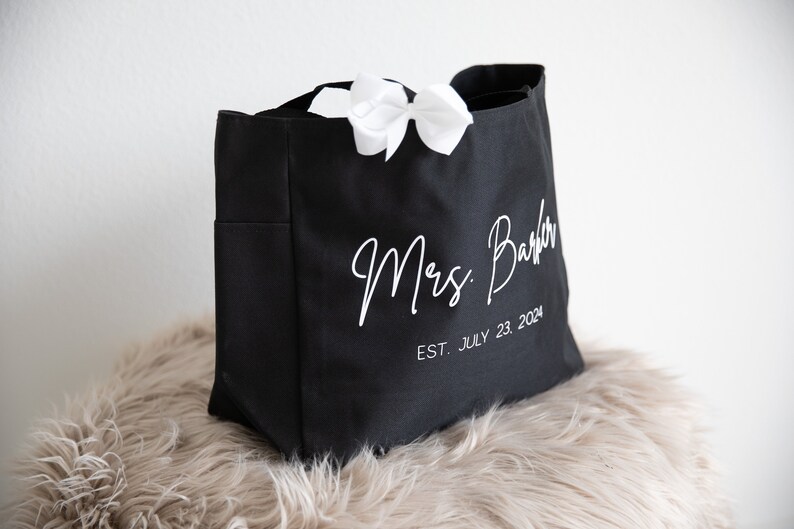 May include: Black canvas tote bag with white text that reads "Mrs.  Barber EST. JULY 23, 2024". A white bow is tied to the handle of the bag.