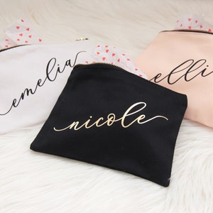 Puede incluir: Tres bolsas de maquillaje personalizadas con letras doradas. Las bolsas son blancas, negras y rosas. Los nombres en las bolsas son "emelia", "nicole" y "ellie".