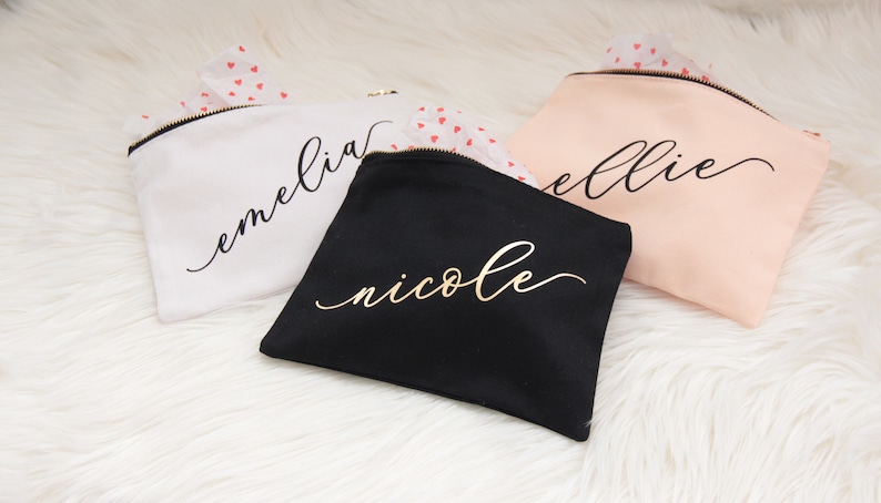 Puede incluir: Tres bolsas de maquillaje personalizadas con letras doradas. Las bolsas son blancas, negras y rosas. Los nombres en las bolsas son "emelia", "nicole" y "ellie".