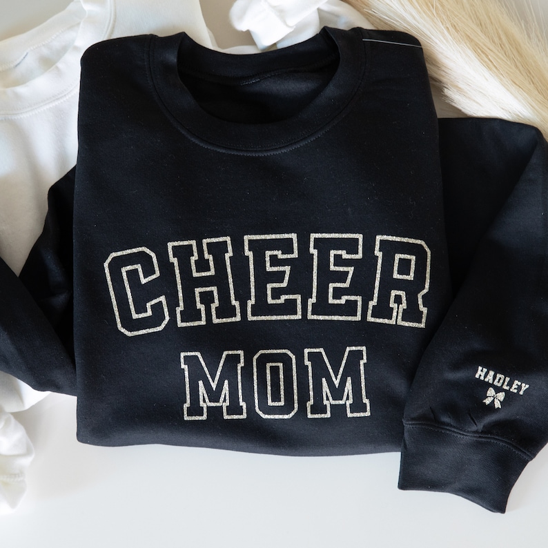 May include: Black sweatshirt with the text "CHEER MOM" in gold glitter lettering. The sweatshirt has a small gold glitter bow with the text "HADLEY" on the sleeve.