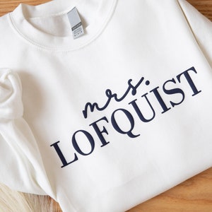 K&ouml;nnte beinhalten: Cremefarbenes Sweatshirt mit dem Text "mrs. LOFQUIST" in Marineblau. Das Kleidungsst&uuml;ck hat einen Rundhalsausschnitt und lange &Auml;rmel. Der Text ist mittig platziert. Der Hintergrund ist eine Holzoberfl&auml;che. Das Sweatshirt scheint aus weichem Stoff zu sein.