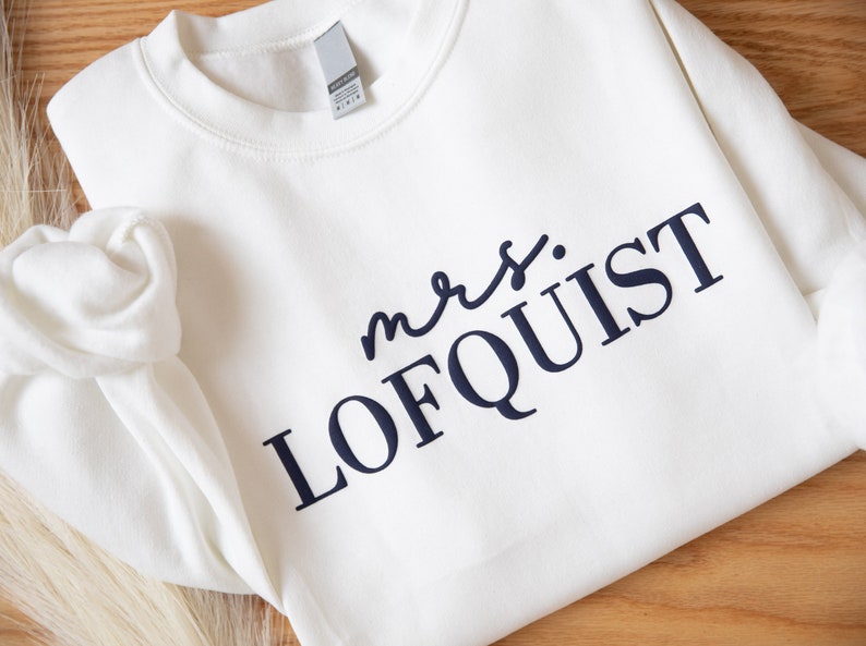 K&ouml;nnte beinhalten: Cremefarbenes Sweatshirt mit dem Text "mrs. LOFQUIST" in Marineblau. Das Kleidungsst&uuml;ck hat einen Rundhalsausschnitt und lange &Auml;rmel. Der Text ist mittig platziert. Der Hintergrund ist eine Holzoberfl&auml;che. Das Sweatshirt scheint aus weichem Stoff zu sein.