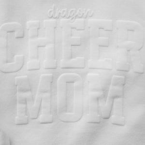 Puede incluir: Sudadera blanca con las palabras "dragon CHEER MOM" en letras en relieve. La palabra "dragon" est&aacute; en cursiva encima de la palabra "CHEER". La sudadera tiene un pu&ntilde;o acanalado.