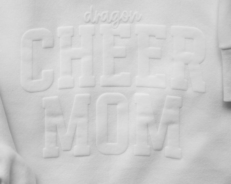 Puede incluir: Sudadera blanca con las palabras "dragon CHEER MOM" en letras en relieve. La palabra "dragon" est&aacute; en cursiva encima de la palabra "CHEER". La sudadera tiene un pu&ntilde;o acanalado.