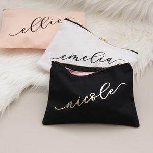 Puede incluir: Tres estuches con cremallera con nombres personalizados en letras doradas. Los estuches son rosa, blanco y negro.