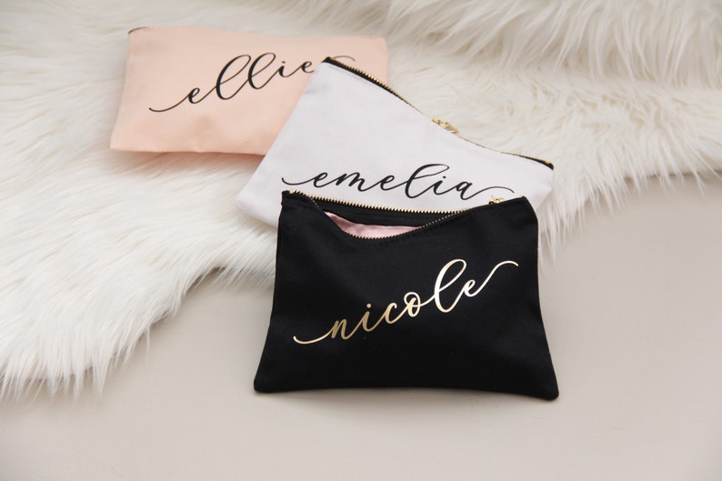 Puede incluir: Tres estuches con cremallera con nombres personalizados en letras doradas. Los estuches son rosa, blanco y negro.