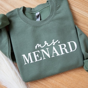 K&ouml;nnte beinhalten: Ein olivgr&uuml;nes Sweatshirt mit dem Text "mrs. MENARD" in Wei&szlig;. Das Sweatshirt ist gefaltet, mit sichtbaren &Auml;rmeln und Kragen. Der Hintergrund ist eine helle Holzoberfl&auml;che.
