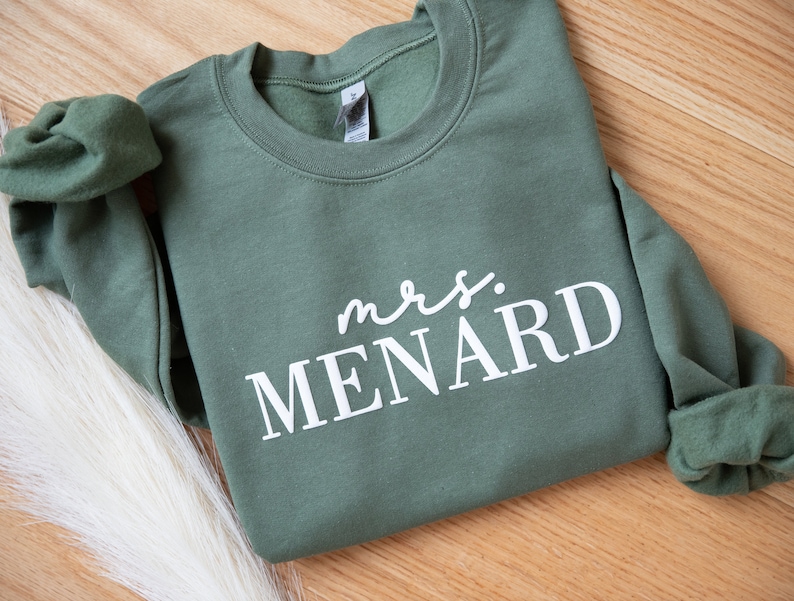 K&ouml;nnte beinhalten: Ein olivgr&uuml;nes Sweatshirt mit dem Text "mrs. MENARD" in Wei&szlig;. Das Sweatshirt ist gefaltet, mit sichtbaren &Auml;rmeln und Kragen. Der Hintergrund ist eine helle Holzoberfl&auml;che.